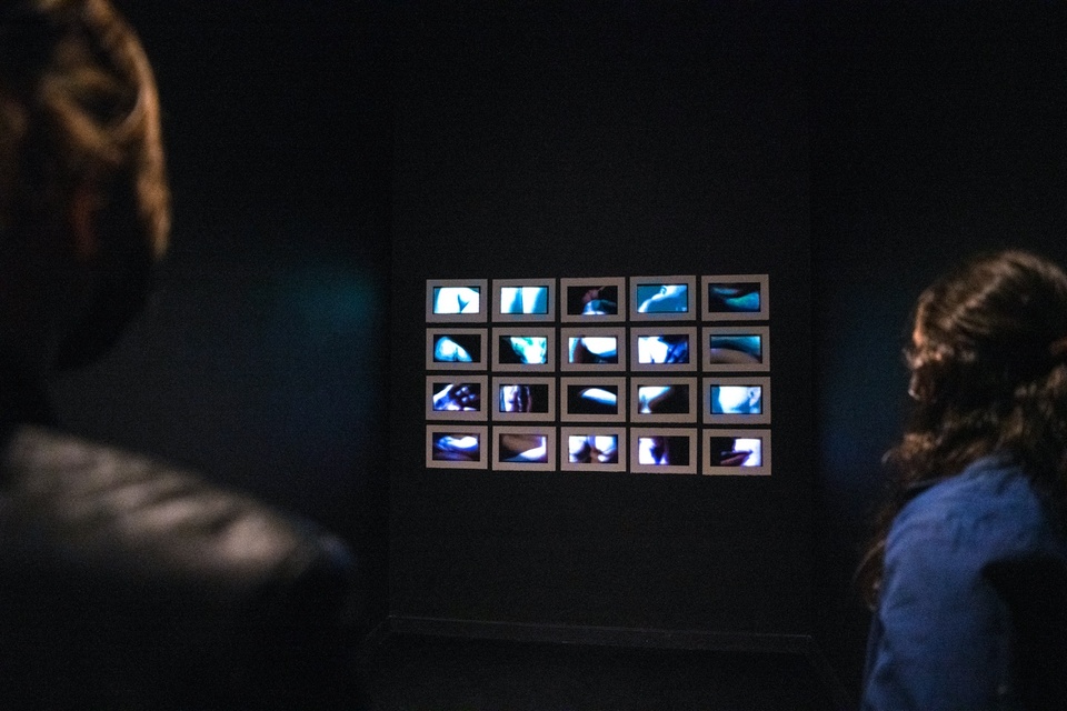 A lightbox in a dark room shows 20 small moving images in a grid. Two figures stand to look at it.