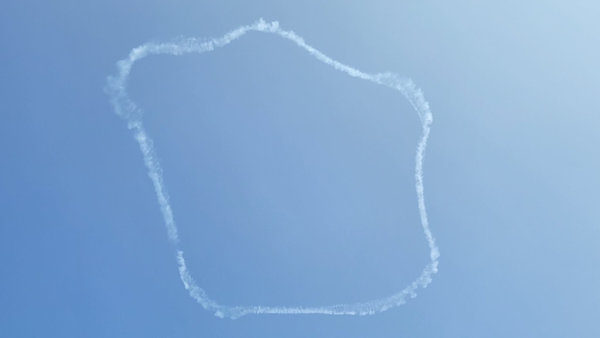 Thin clouds created by a skywriting airplane delimit a roughly square area in the middle of a blue sky, seen from directly below.