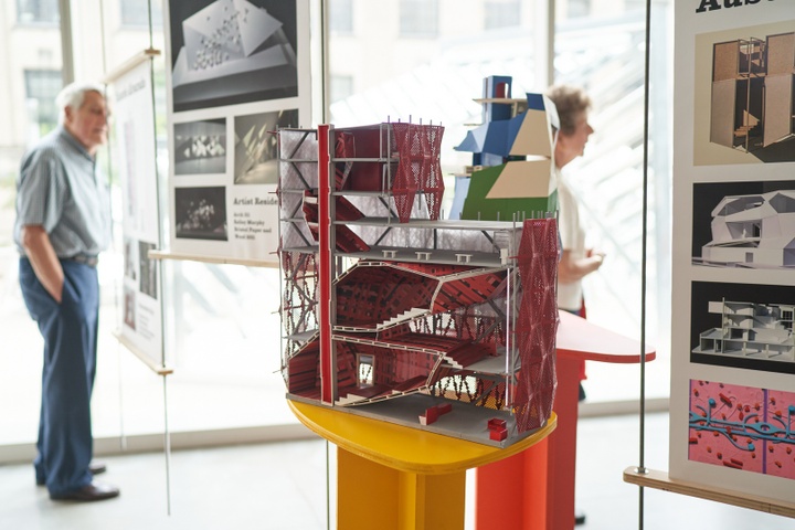 Closeup of a colorful model cut open in section, displayed on a yellow stand. The building is 6 floors. The bottom two floors contain auditorium spaces with red walls and accents. The building features a red lattice envelope around the exterior.