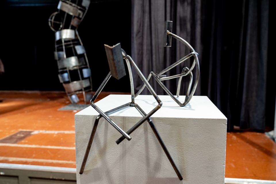 Two abstract metal stick men crouching on a white pedestal.