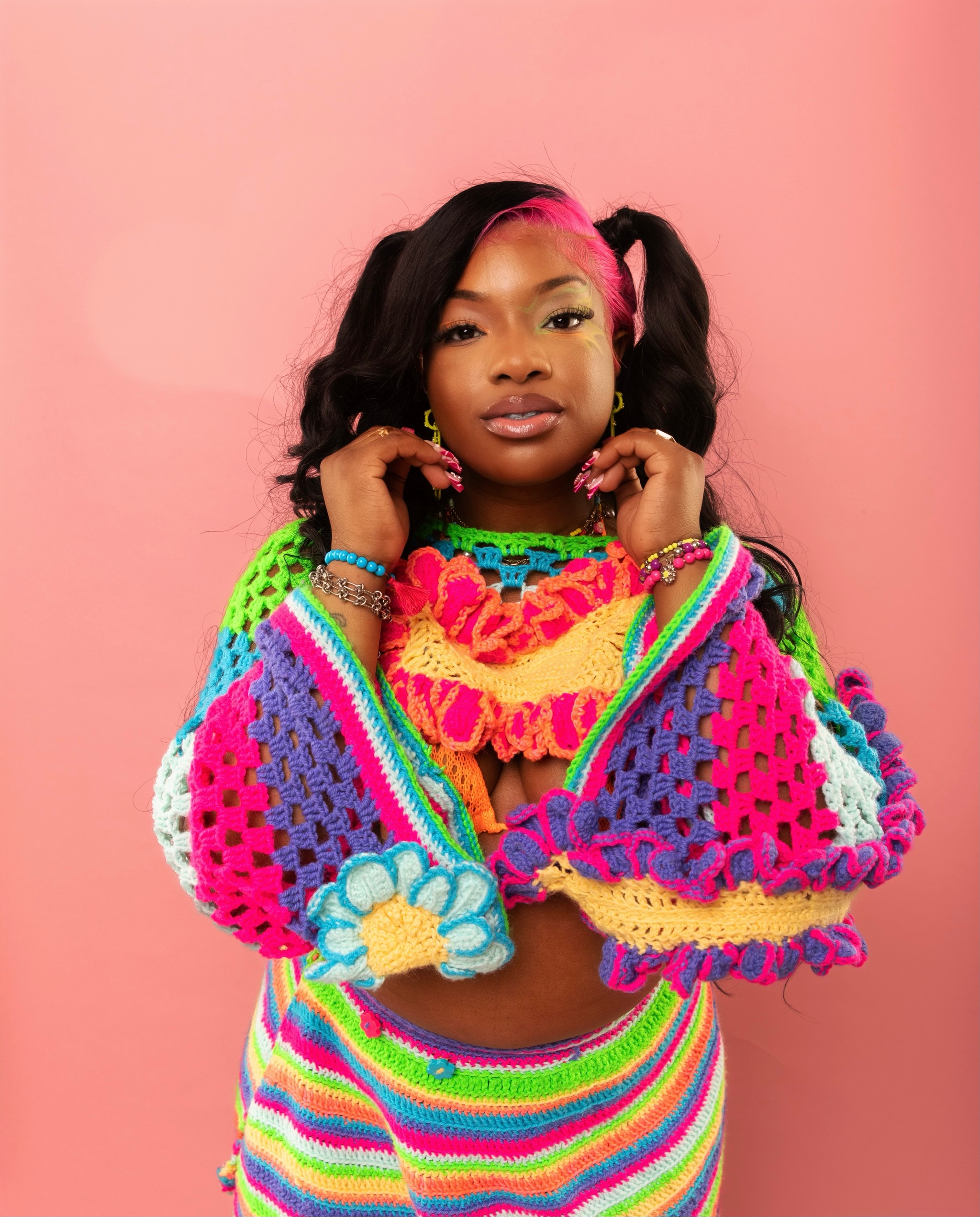 A Black woman wearing brightly colored skirt and crocheted top stands against a pink wall. She holds her hands up to either side of her face. She wears her hair in long pigtails and has a streak of bright pink hair on the left side of her head. 