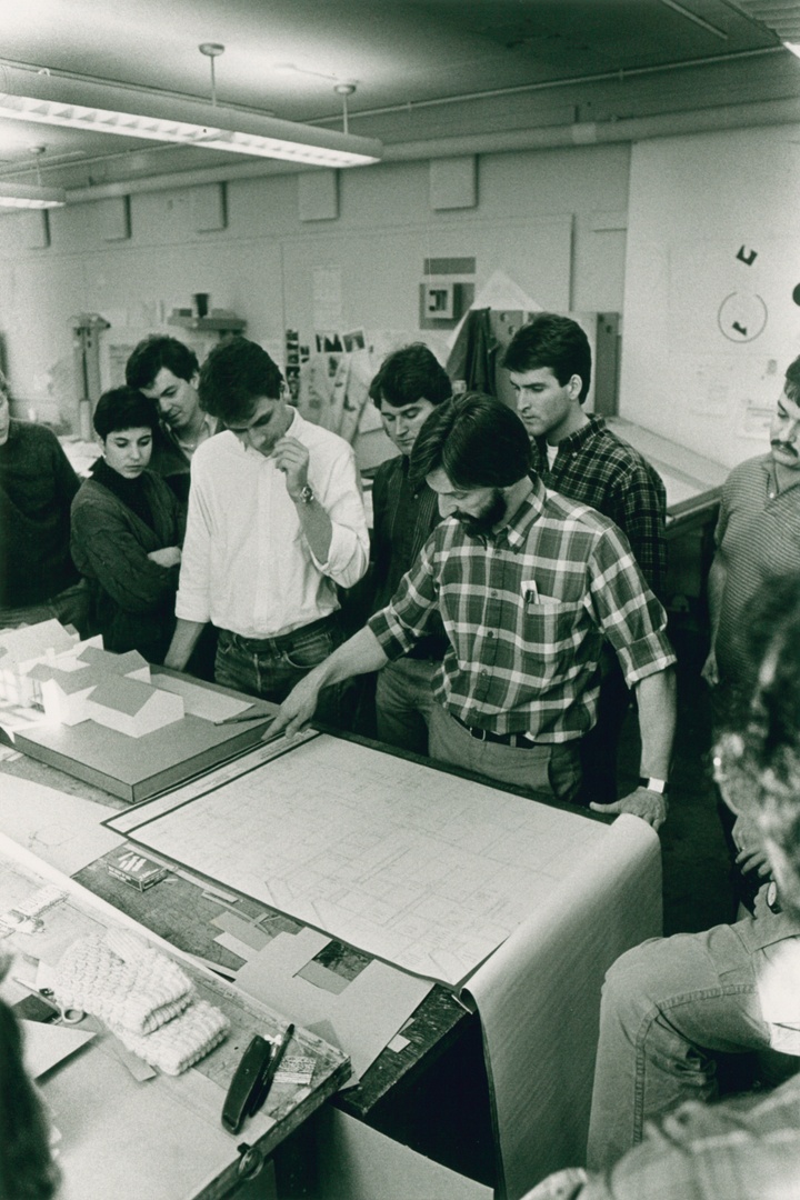 Black and white photo of a person gesturing to a blueprint document on a studio table while others look on.