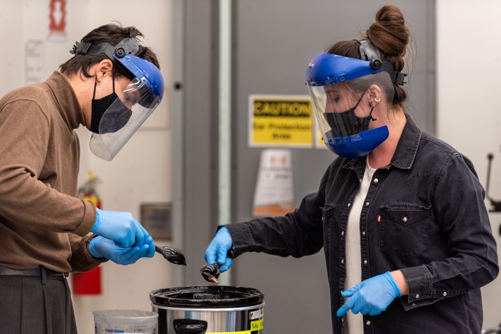 A student and an instructor wearing face shields and gloves use plastic tongs to dip something out of a crockpot.
