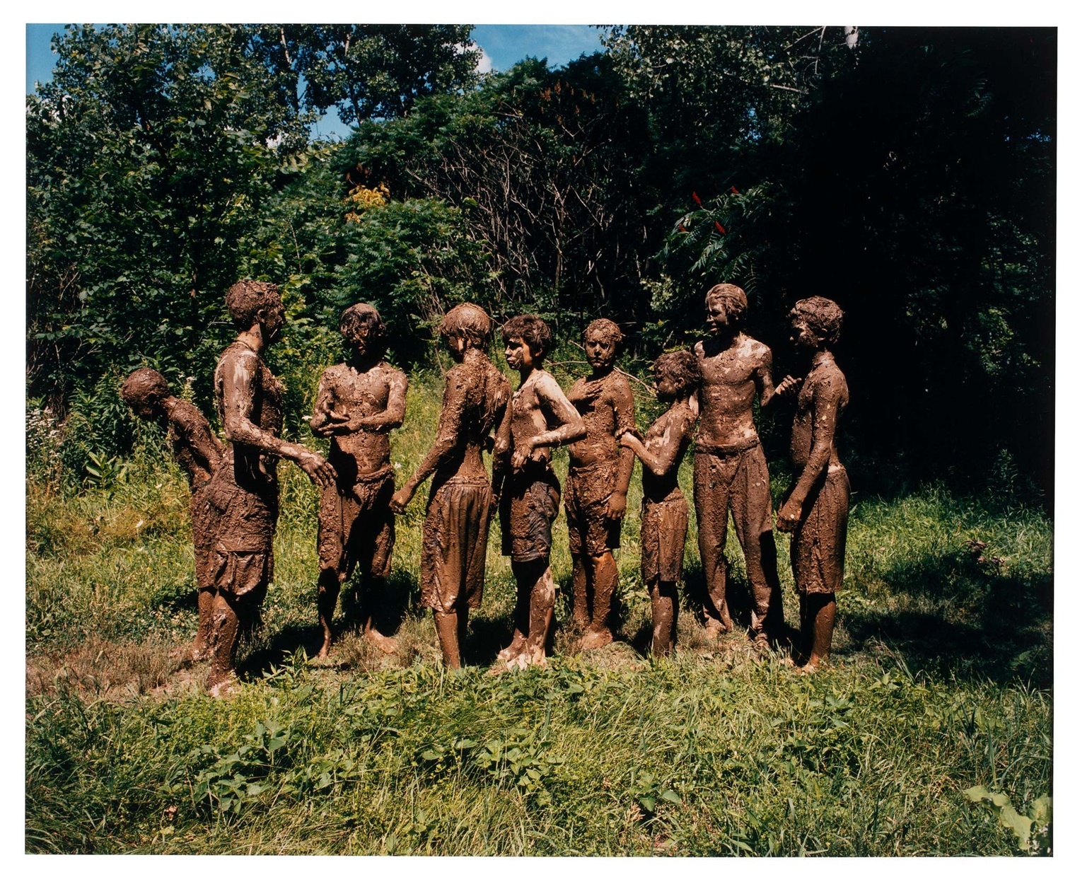 Nine mud-covered boys stand in a line on grass with lush trees in the background.