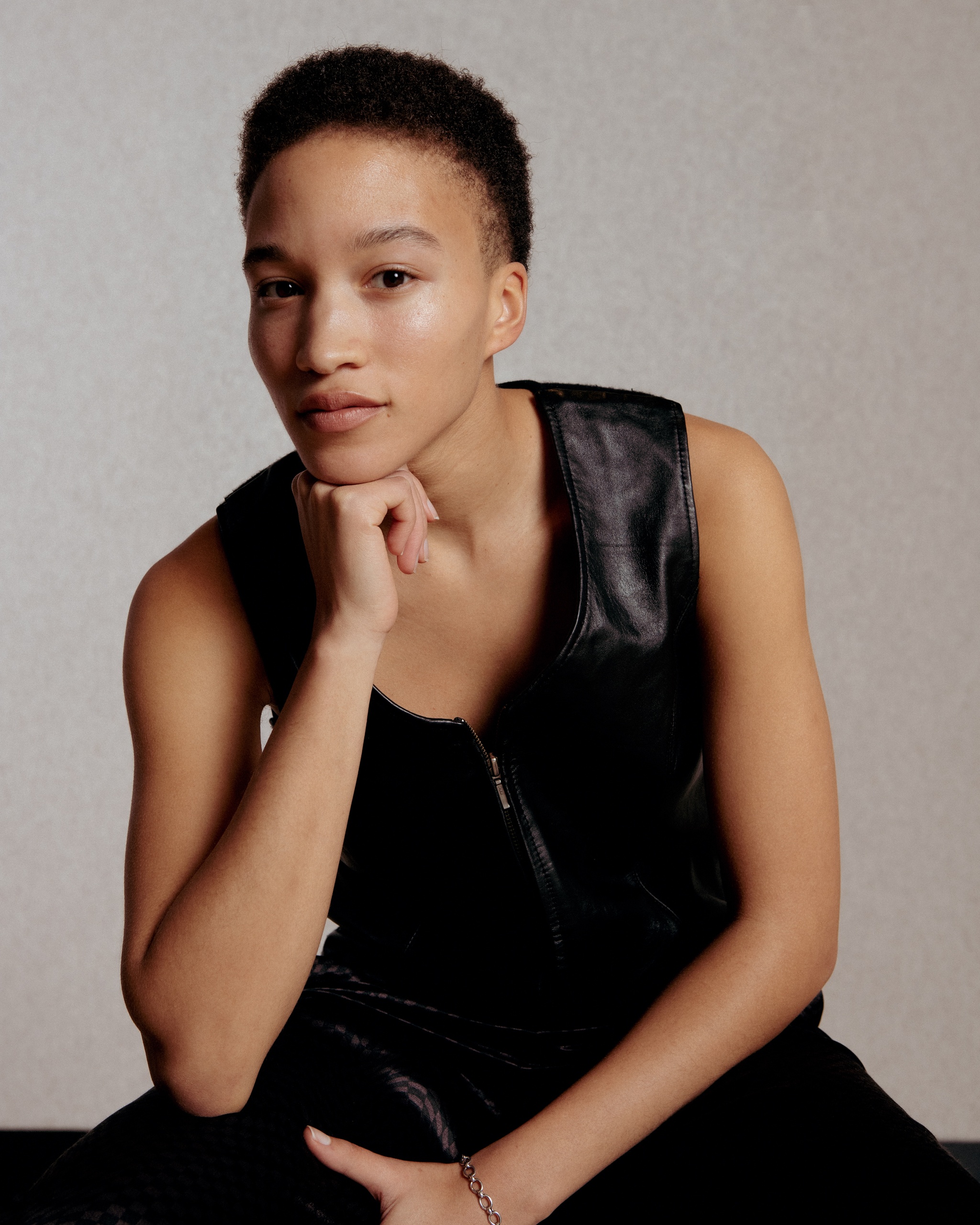A portrait of artist Asia Stewart, a Black woman with light brown skin who poses with her elbow on her knee and her chin supported by a loose fist. She looks directly at us from a three quarter profile with one eyebrow slightly raised. Asia has short hair and wears a black leather vest. Photo by Dana Golan