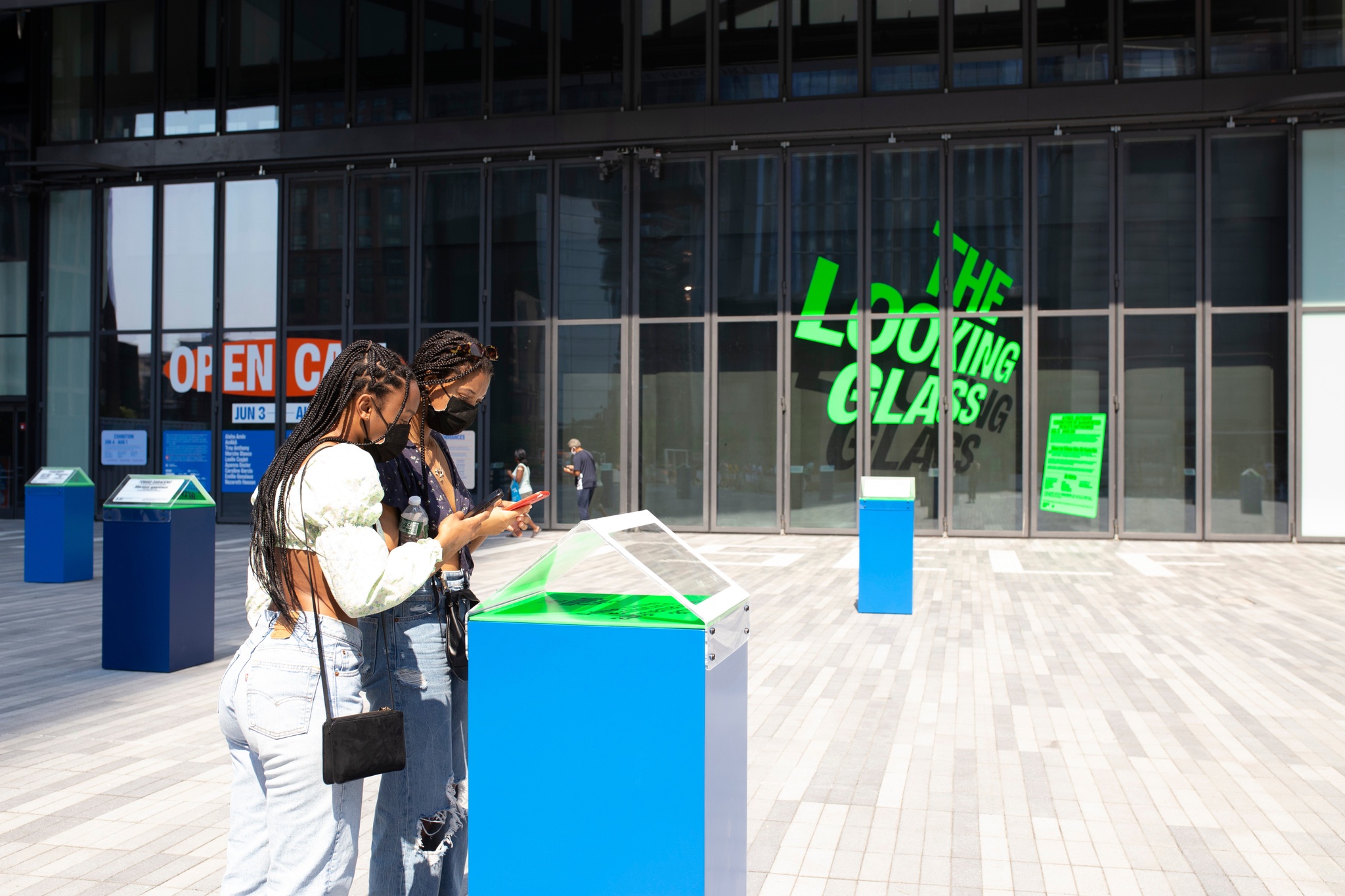 Two people with long braids lean over a bright blue plinth used to activate the exhibition The Looking Glass on a sunny outdoor plaza