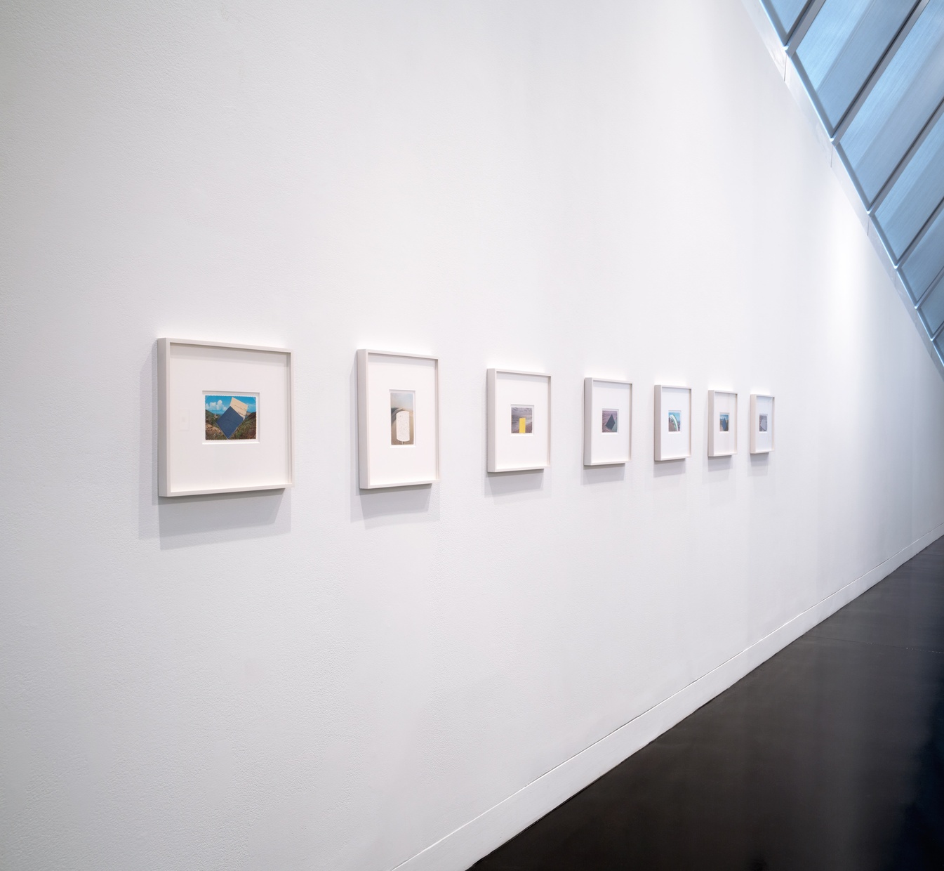 Seen from an angle, a row of framed postcard collages hangs on a white wall.