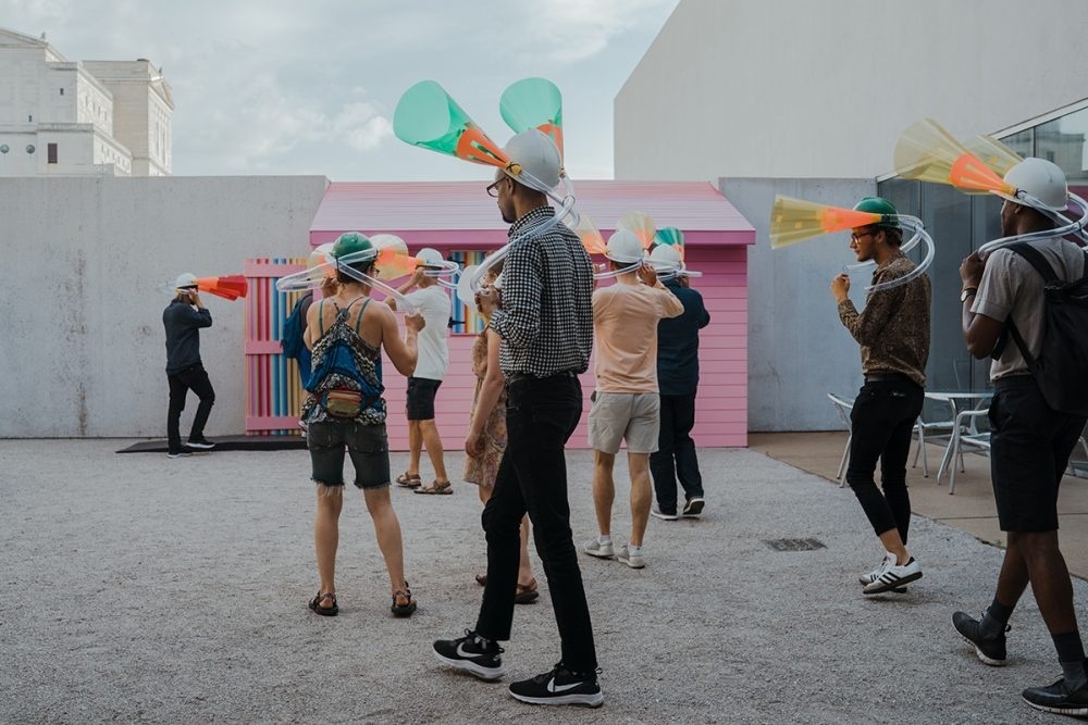 Group of people wearing cylindrical sound pieces as headwear approaching a pink colored house installation at the Contemporary Art Museum