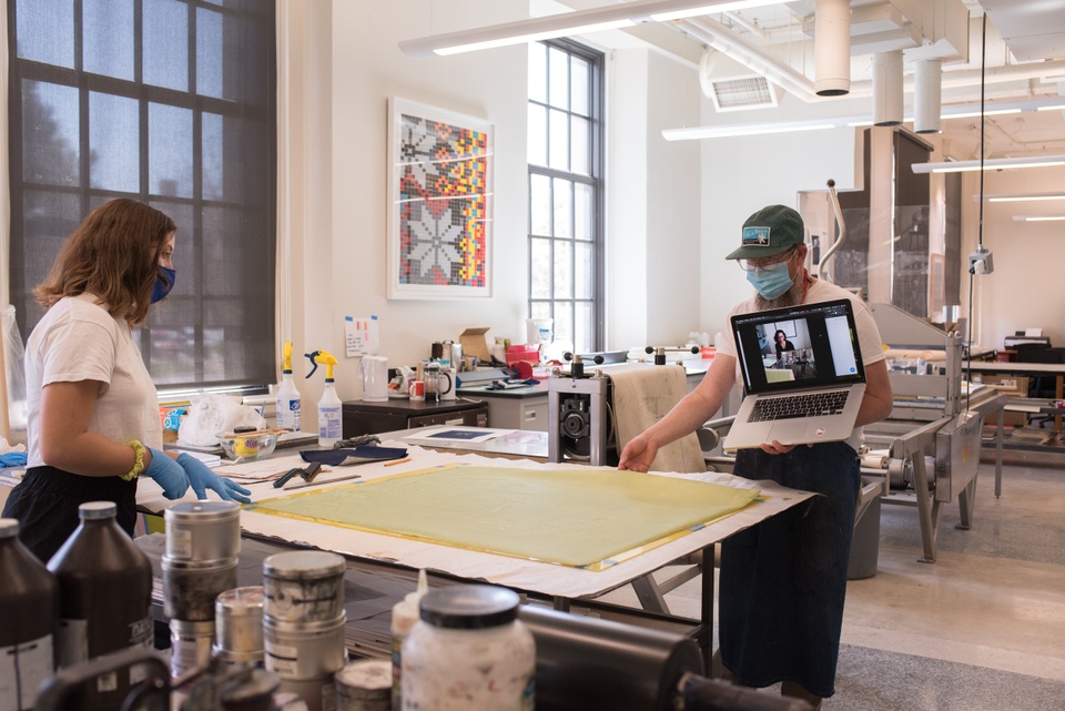 Person holds a laptop with video chat up to view a large cyanotype print on the table of a printmaking studio.