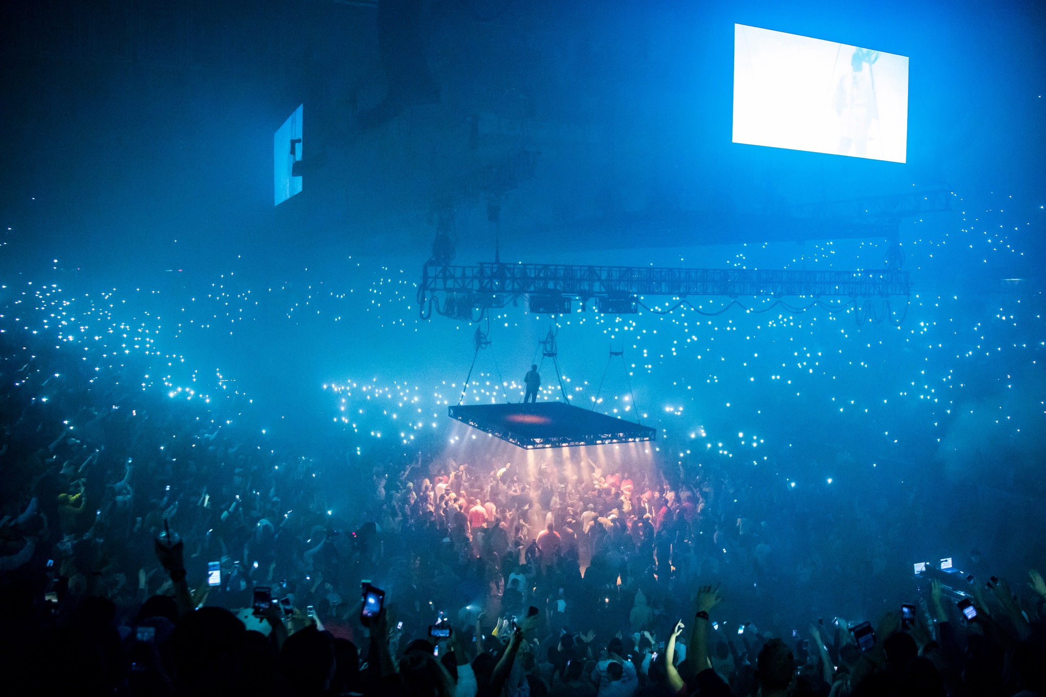 A crowd at an arena concert watching a performer on a raised platform at the center