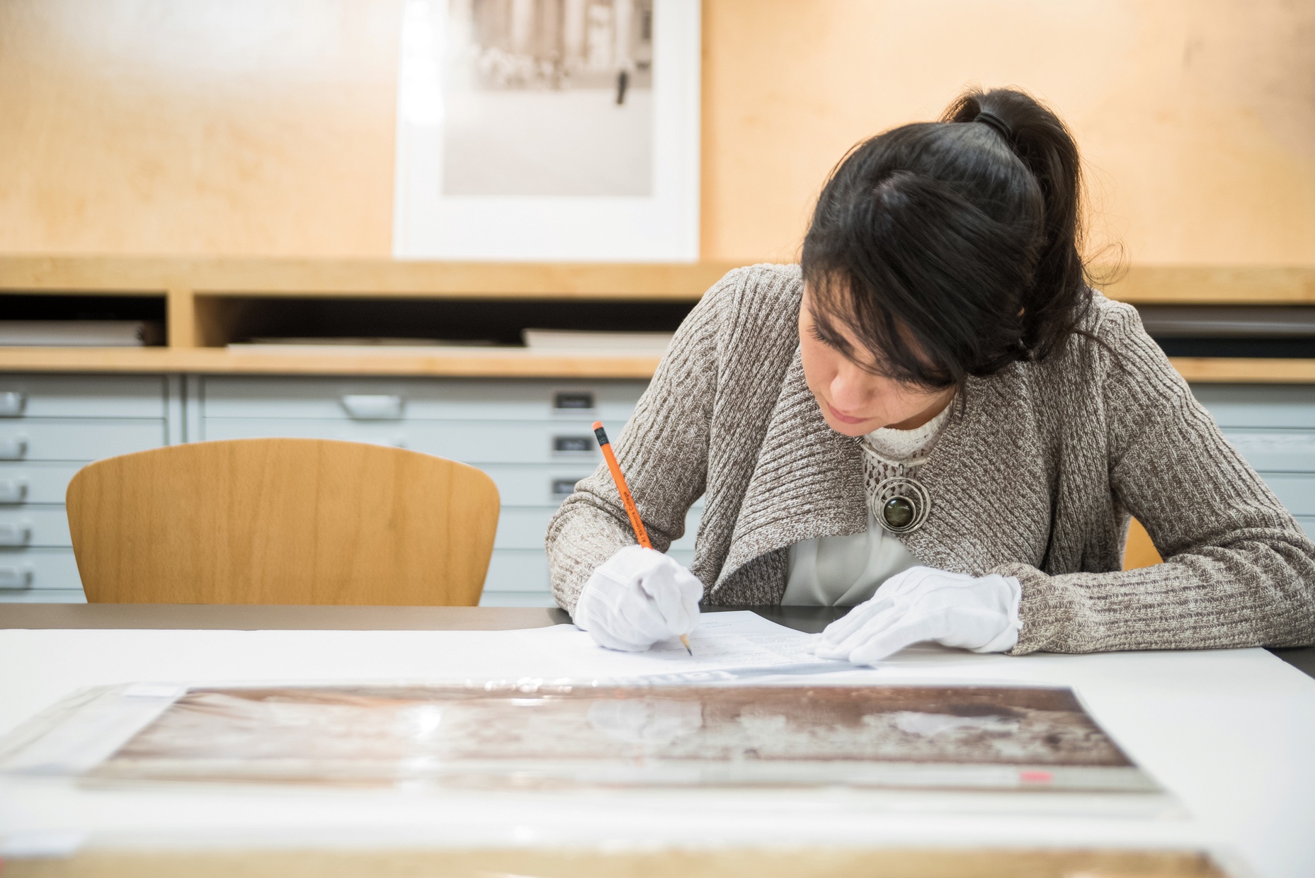 Ana-Joel Falcon-Wiebe, a light-skinned female, takes notes with a pencil on a piece in the museum's collection while wearing white gloves.