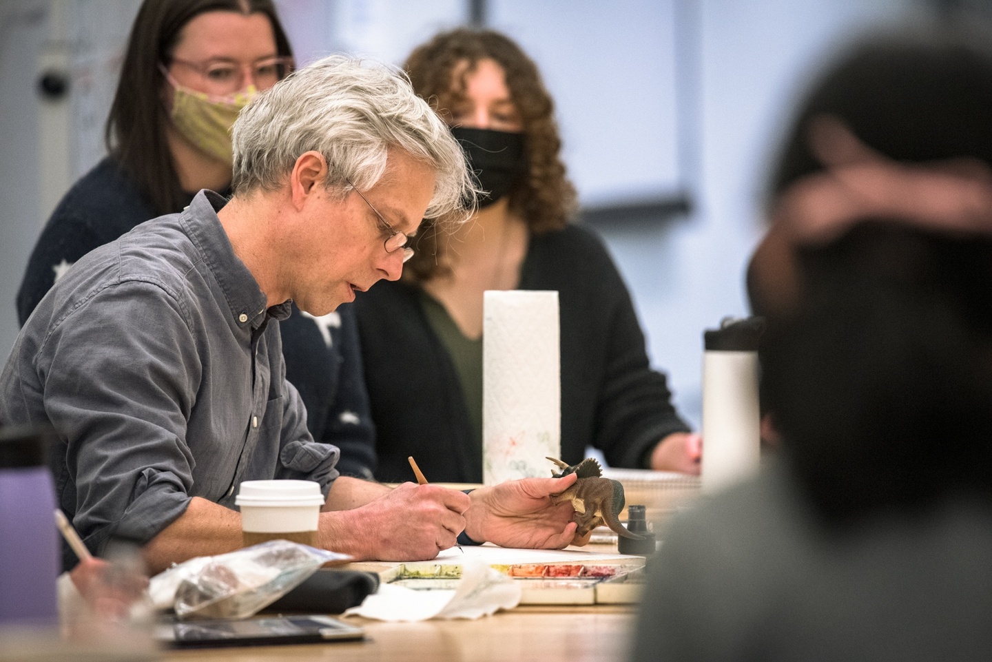 Person works on a watercolor painting at a table while holding a plastic dinosaur model in the off-hand. A group of students watches.