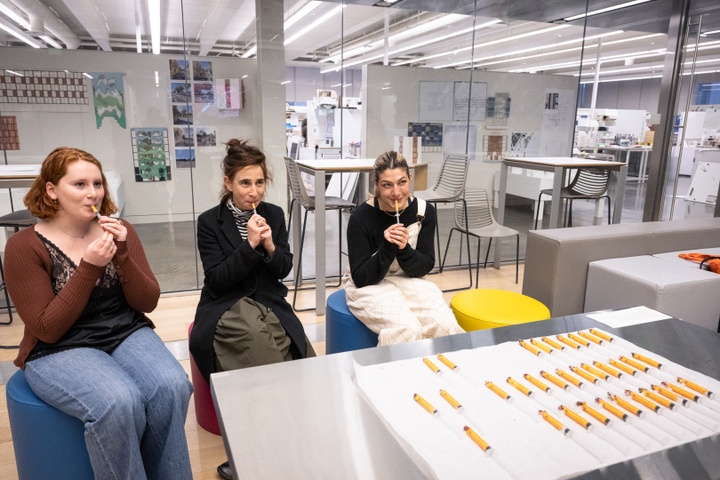 Three people sit by a table covered in syringes and inject a yellow paste into their mouths.