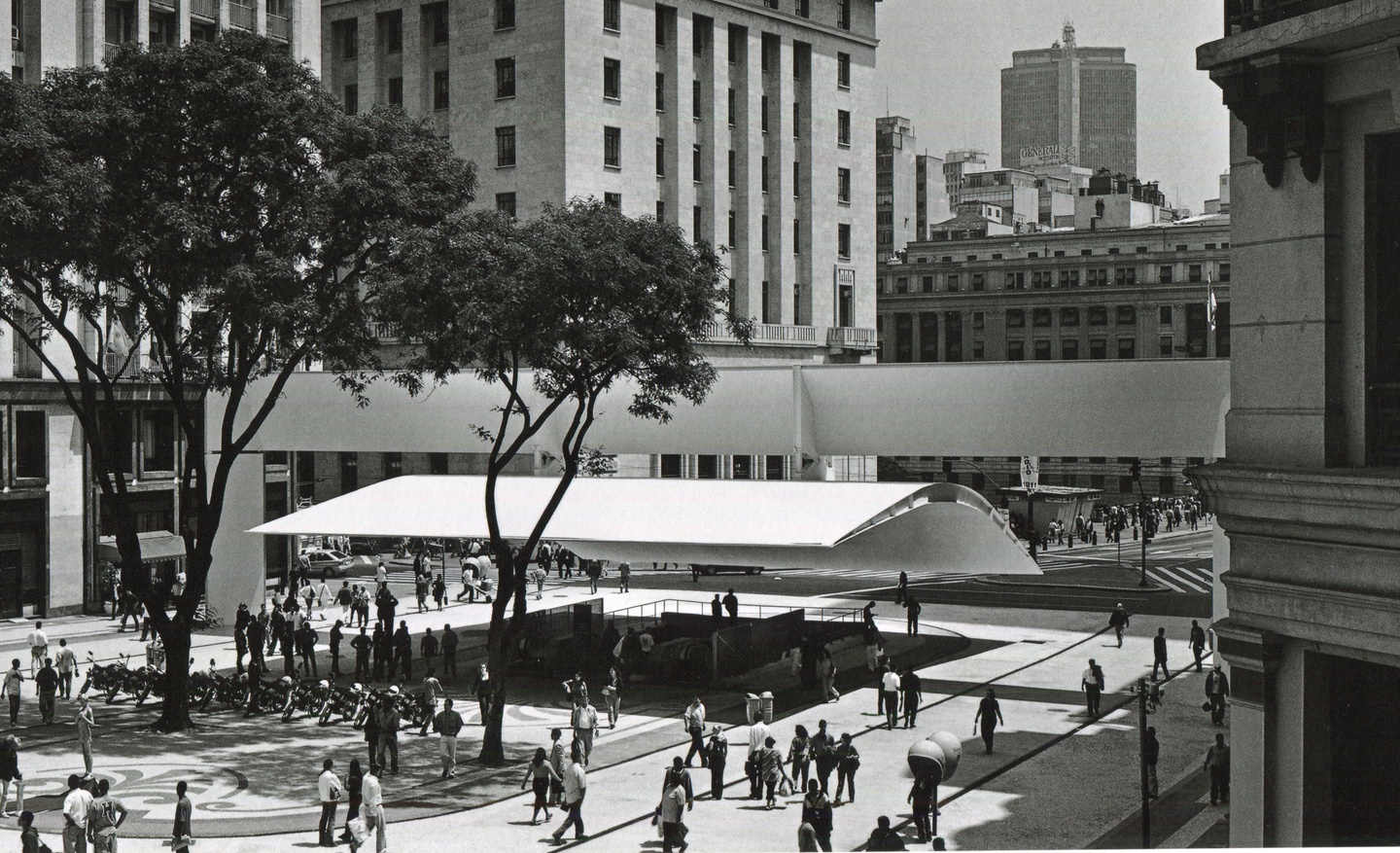 Black-and-white photo of people in a spacious city plaza, with a couple of tall trees and a canopy overhang