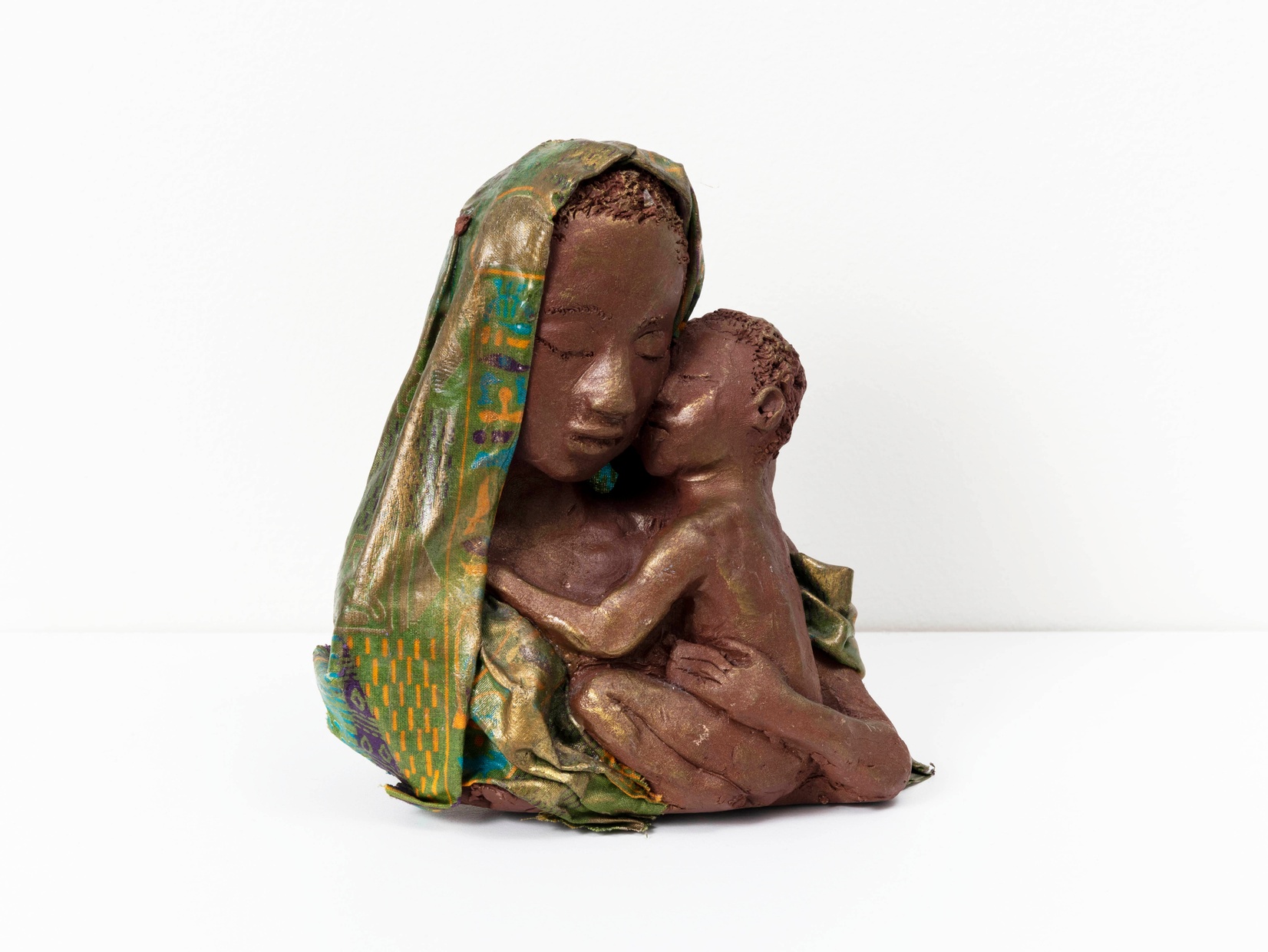 A dark clay bust of a woman wearing a green headscarf and holding a baby in her arms up to her cheek.