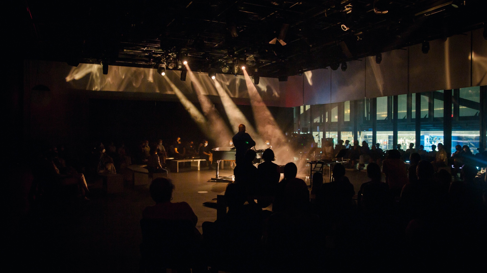 The artist Justin Hicks whose face and features are obscured stands at the center of an audience surrounding him. The space is stands in is lit dramatically by stage lights that send beams of light through the foggy air.