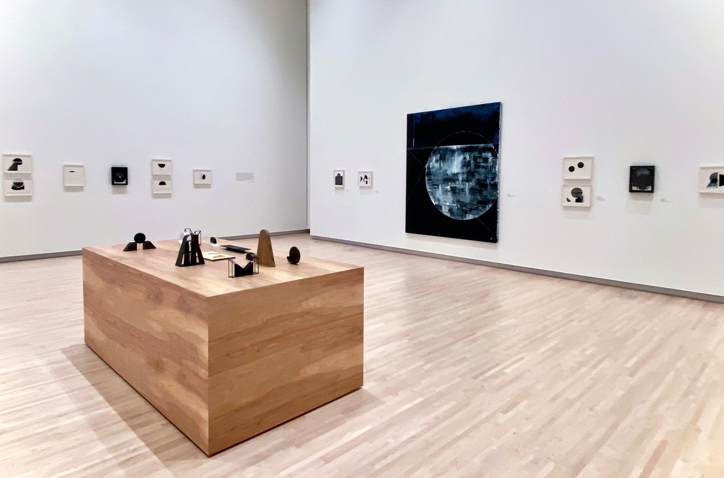 An exhibition installation with smaller geometric drawings, a large, dark geometric painting, and a wooden table with small geometric sculptures
