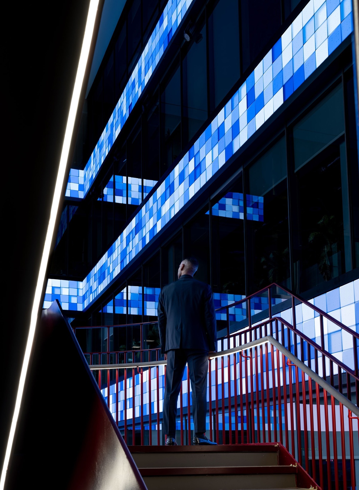Cinematic photograph of businessman standing on stairwell, looking out at blue, geometric graphics displayed on LED screens