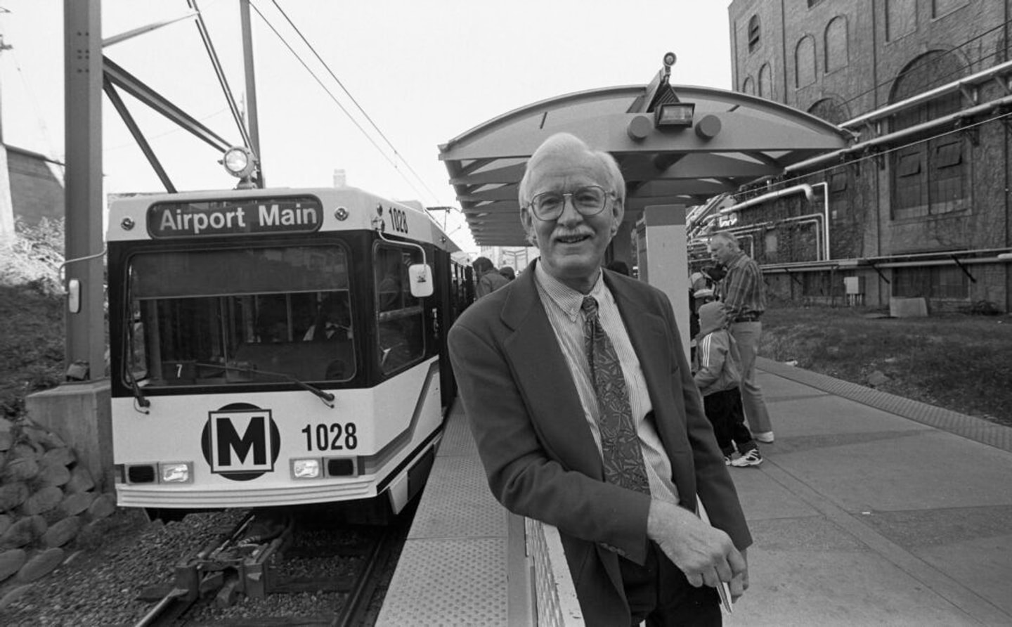 A black and white photo of a man leaning against a railing with a lightrail car behind him.