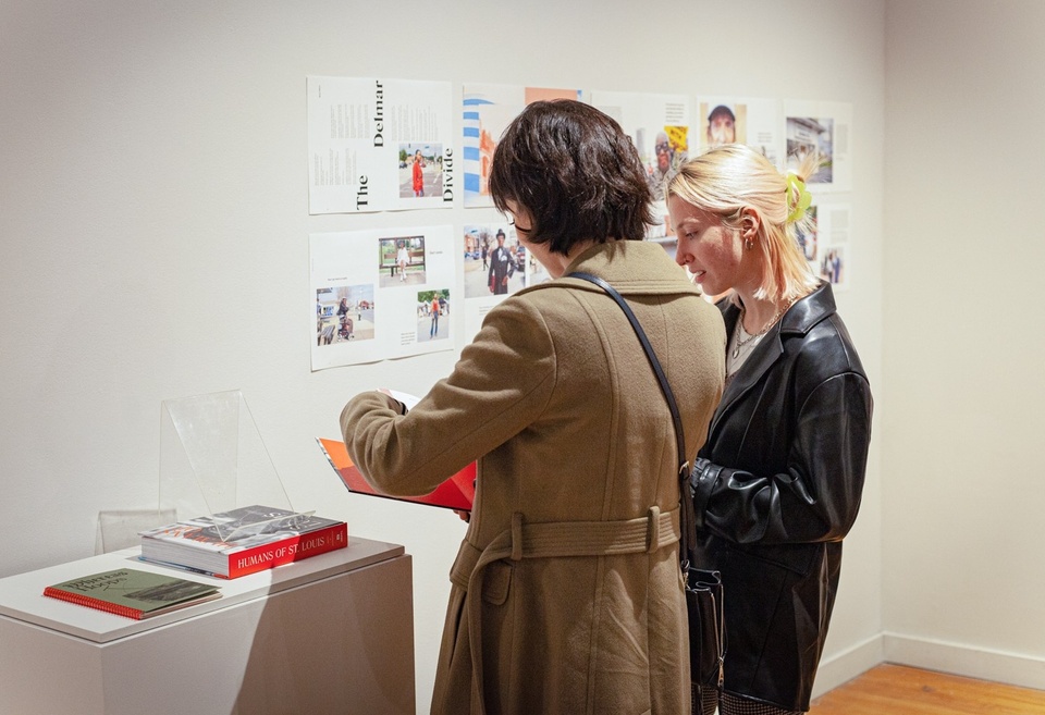 Two students look at a copy of the "Humans of St. Louis" photobook. On the wall behind them are prints of several page layouts from the book.