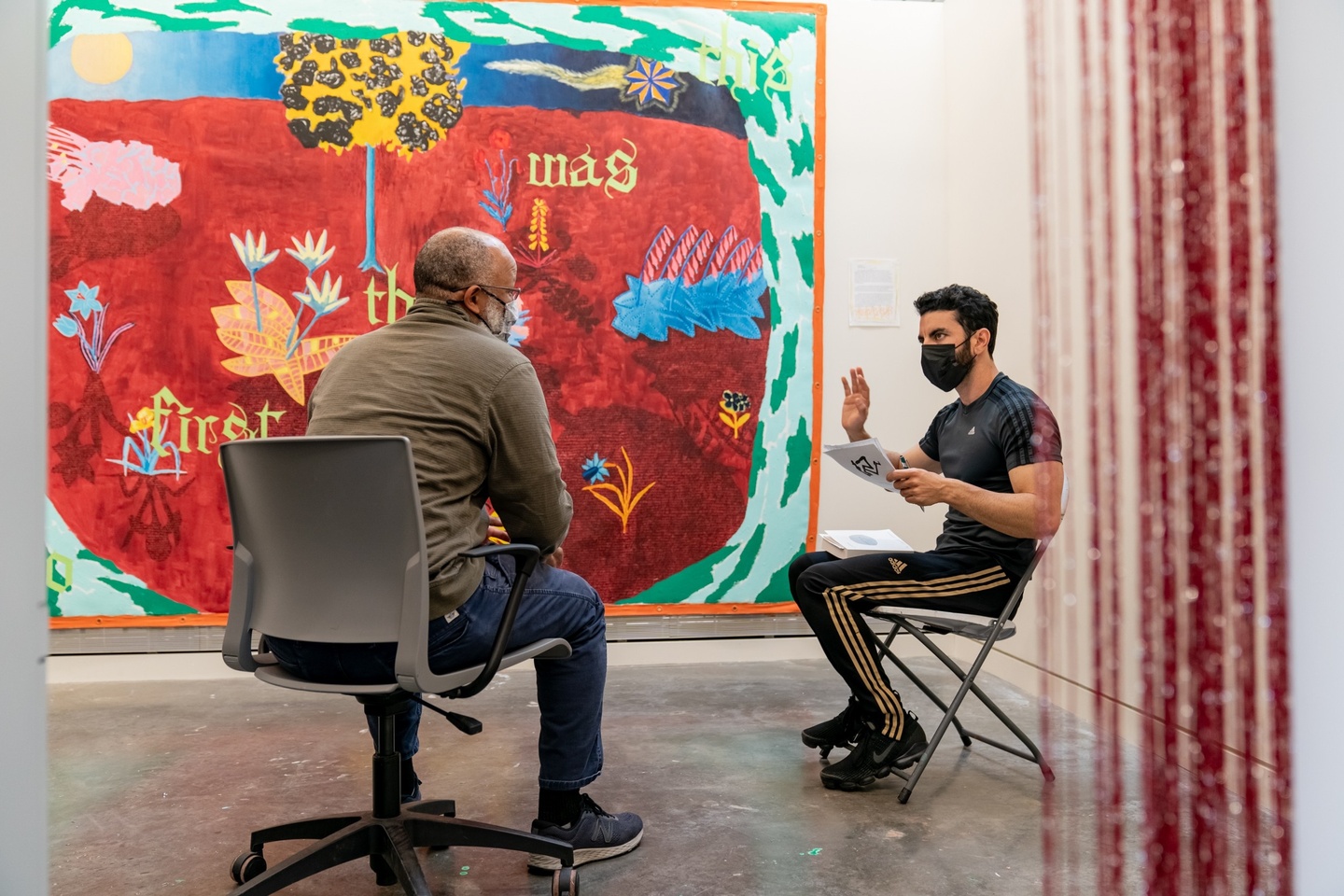 A student and faculty member sit in the student's studio, where a large, colorful artwork hangs on the wall.