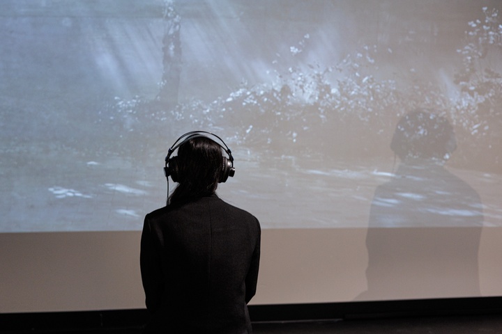the artist with her back to us wearing headphones standing in front of a large screen