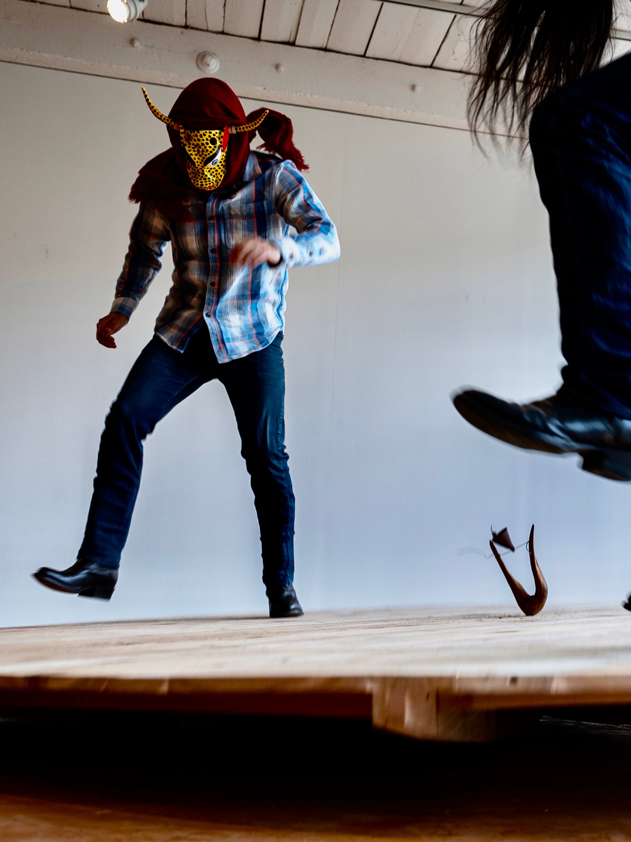 Two performers stand in a nondescript room on a wooden platform. They are both seen from an angle slightly below, one of them is cut off by the frame of the image so we see only legs and long hair hanging down from where the performer bends over. The other performer is seen in full, wearing jeans and boots, a blue plaid shirt, and a head covering including a ceremonial mask that resembles a jaguar’s face with two long, thin horns. Both are suspended in air slightly above the ground, stomping.