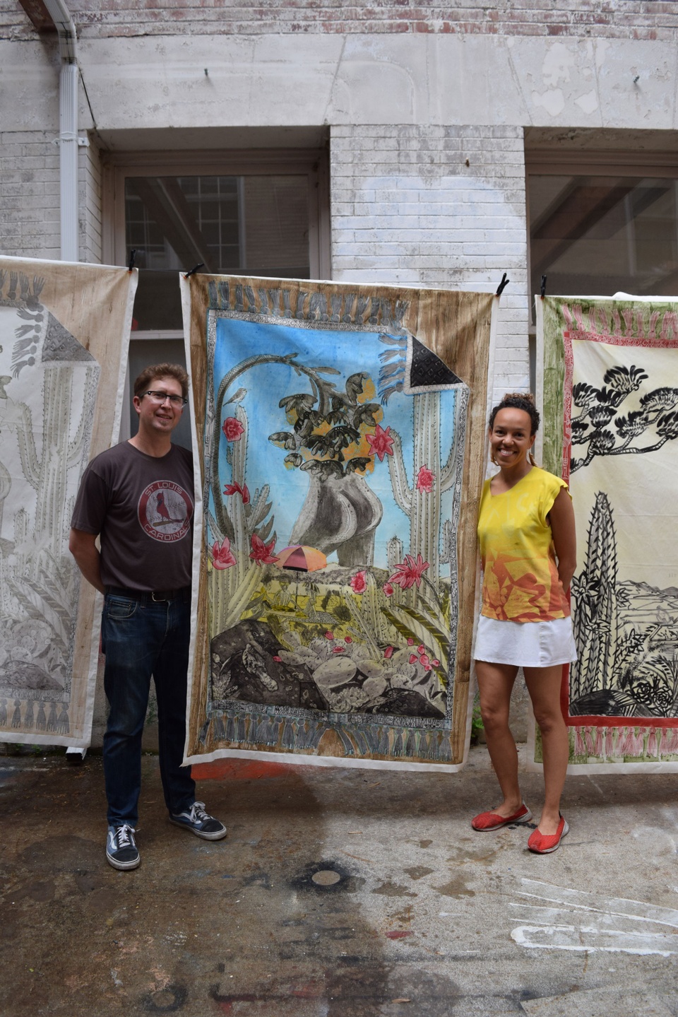 Paula Wilson and Tom Reed flank her large print that is taller than them both and features flowers, cacti, a large butt and people sitting under an umbrella