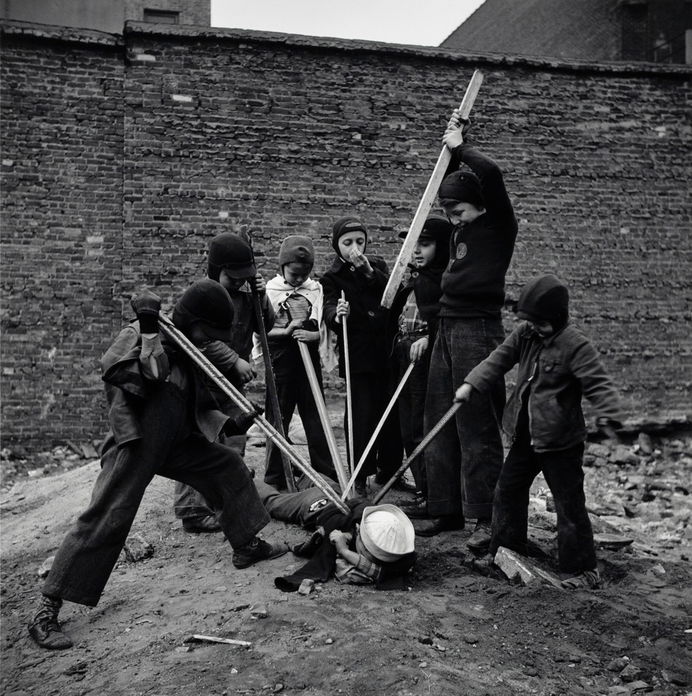 A black and white photograph of a group of six children pressing sticks onto a boy lying face down on a dirt ground while another boy raises a large stick above his head towards the boy on the ground.