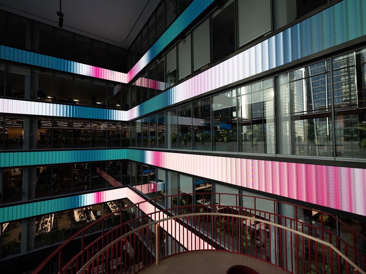 Photograph of final experience design of atrium from circular stairwell, of four long horizontal rows of LED screens in white, pink and turquoise gradients punctuated between four floors