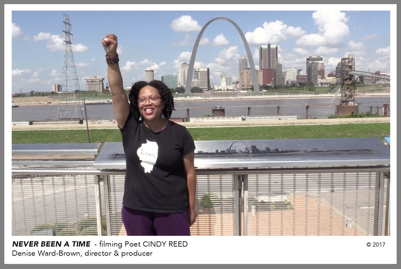 Video still of a poet standing outside smiling, right arm raised and her hand formed in a fist, with the riverfront and St. Louis Gateway Arch in the background.