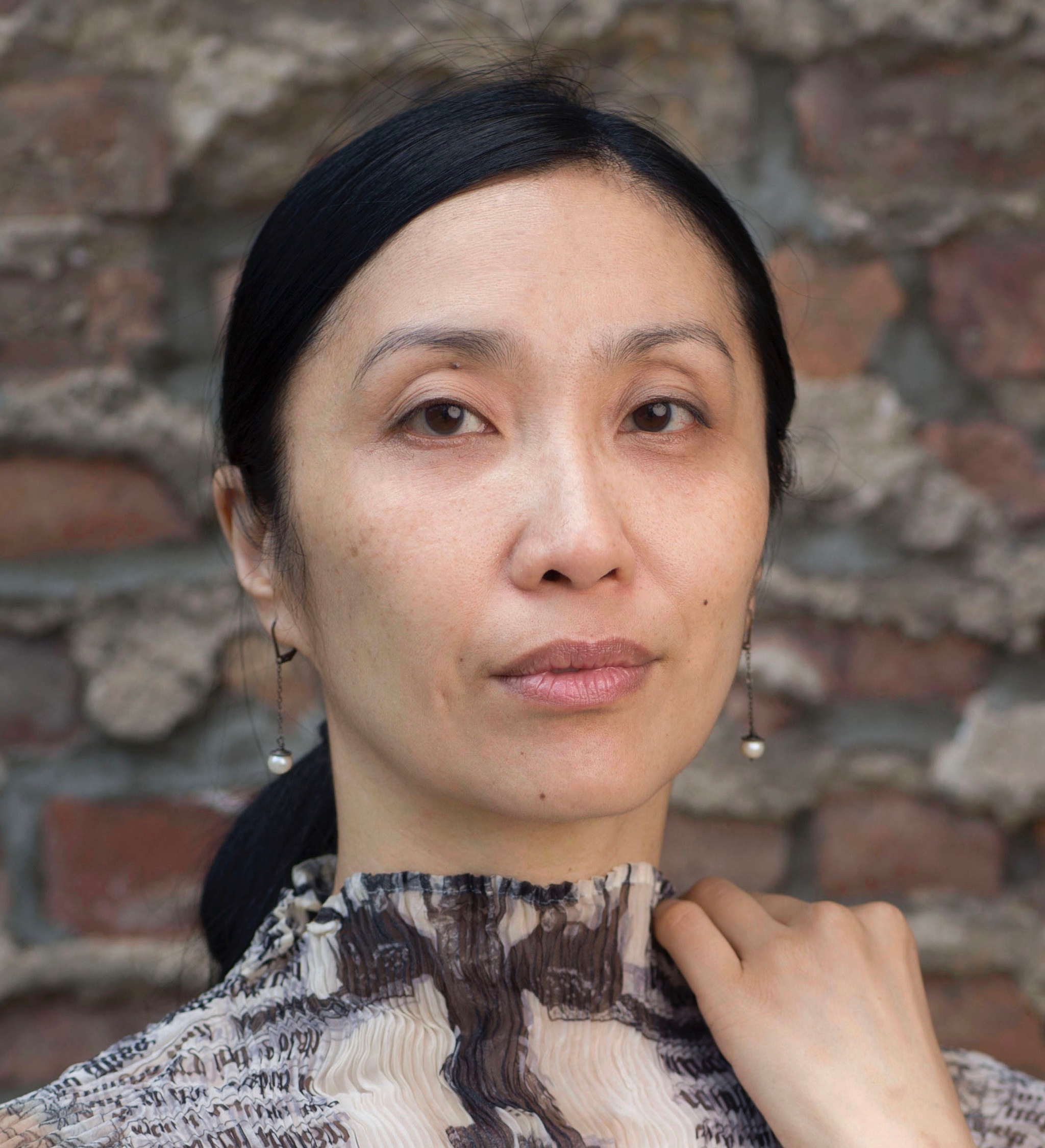 A headshot of Janet Wong, an Asian woman who poses against a brown brick wall. Her dark hair is pulled back behind her head. She holds one hand to her shoulder and looks intently at us. Photo by Eric Politzer.