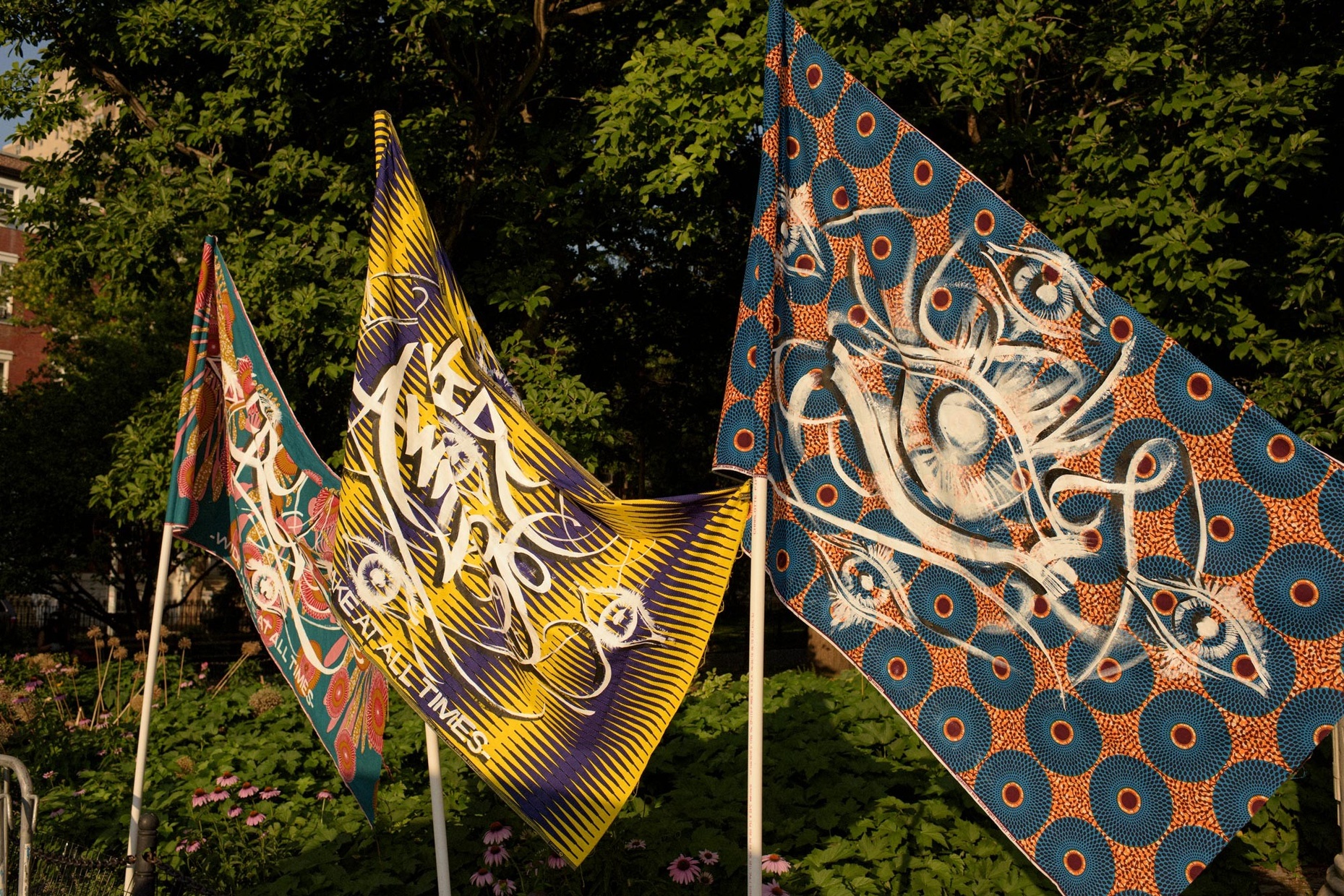 A photograph of three colorful flags blowing in the wind, with green leaves behind them. The flag on the right bears five white eyes atop a blue and orange pattern.