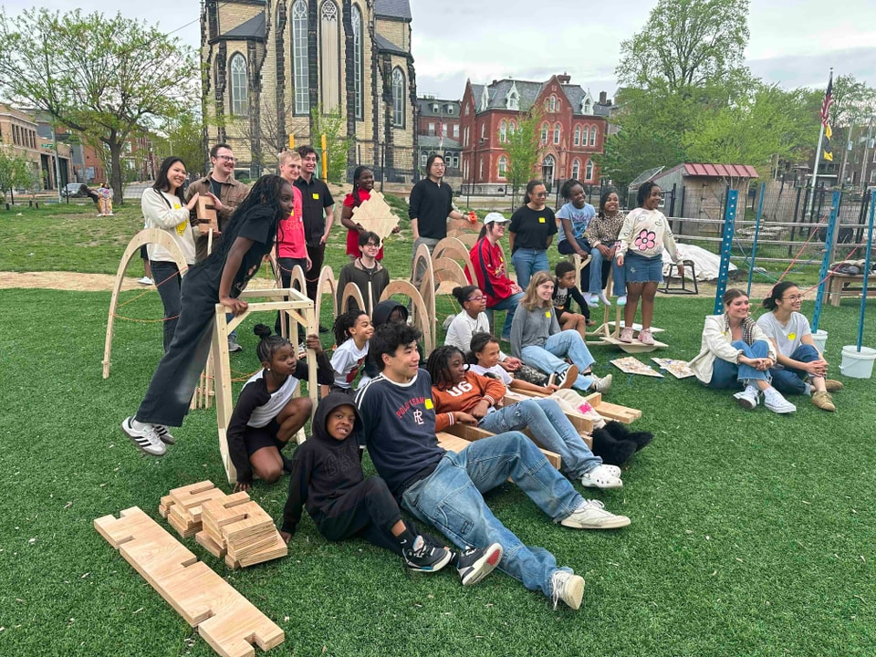 A large group of students and children pose outdoors with modular wooden play structures on a lawn in front of a historic stone church.