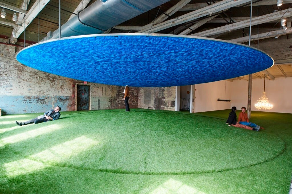 On a green hill in a gallery, people sit and stand, relaxing, as if in a park. A blue yarn inverted-hill form floats above it. The head of a person standing beneath the form disappears into the space above.