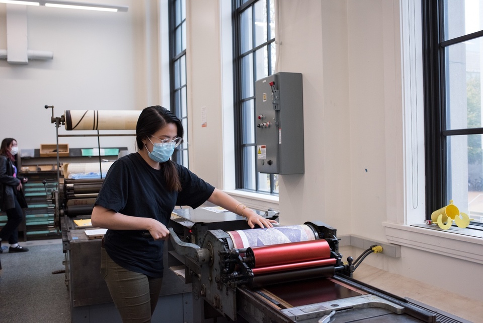 Person cranks the handle on a roller and runs a print through it.