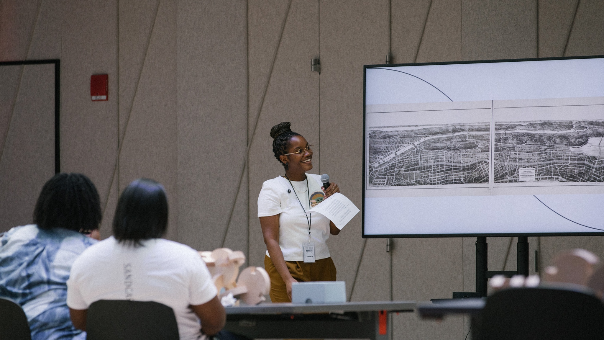 The artist Ladi'Sasha Jones stands beside a video screen in front of an audience seated at tables. On the screen is a 19th century map of Harlem and New York City. 