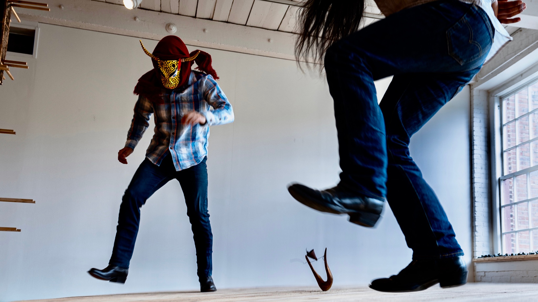 Two performers stand in a nondescript room on a wooden platform. They are both seen from an angle slightly below, one of them is cut off by the frame of the image so we see only legs and long hair hanging down from where the performer bends over. The other performer is seen in full, wearing jeans and boots, a blue plaid shirt, and a head covering including a ceremonial mask that resembles a jaguar’s face with two long, thin horns. Both are suspended in air slightly above the ground, stomping.