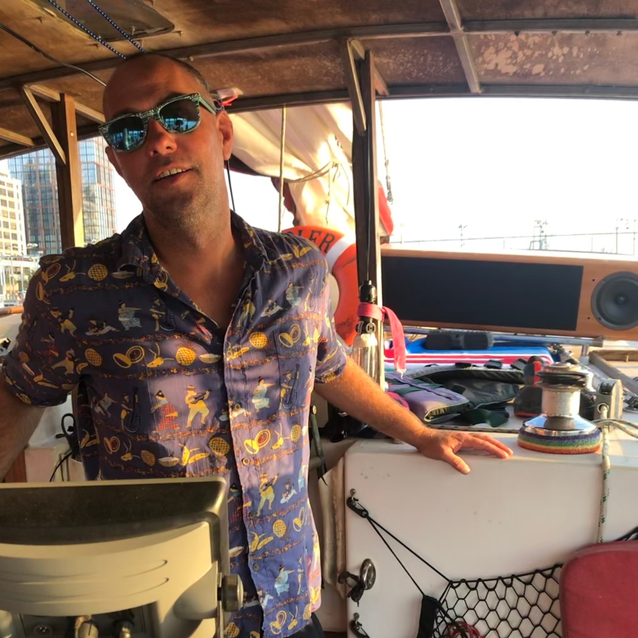 A man stands in sunglasses and a shirt with a colorful, cartoonish print within a sail boat