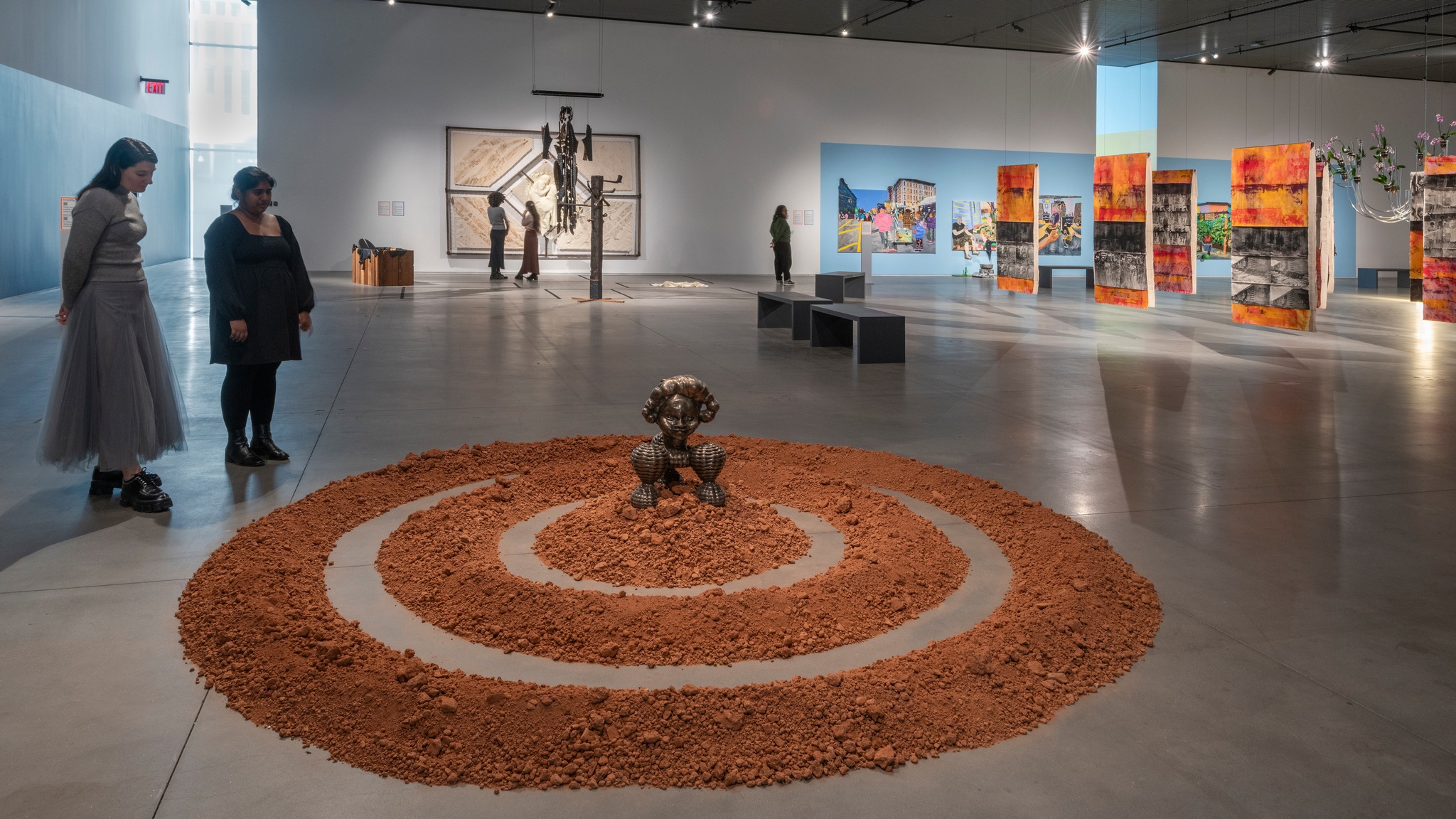 Two gallery visitors stand in an art gallery looking down at a wide artwork on the floor. It consists of three concentric rings of red dirt. At the center sits a bronze sculpture inspired by the Benin Bronzes, historical artworks from Benin, present-day Nigeria. In the background are other paintings and installations.