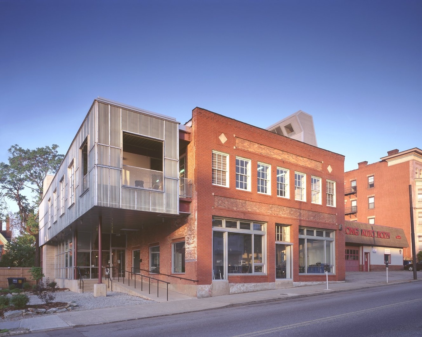 A perspective photograph of a brick building along the street with a steel double façade on the upper level, cantilevered over a ramp way.