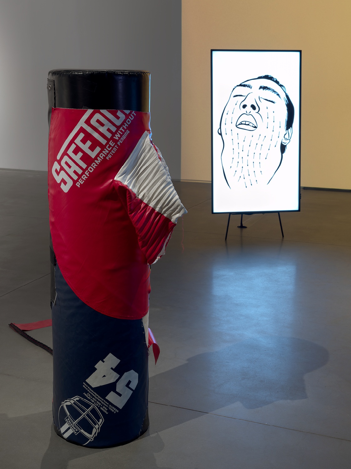 A tackling dummy that has been restitched stands upright with a vertical video monitor in the background that depicts a drawing of a man's face turned upward