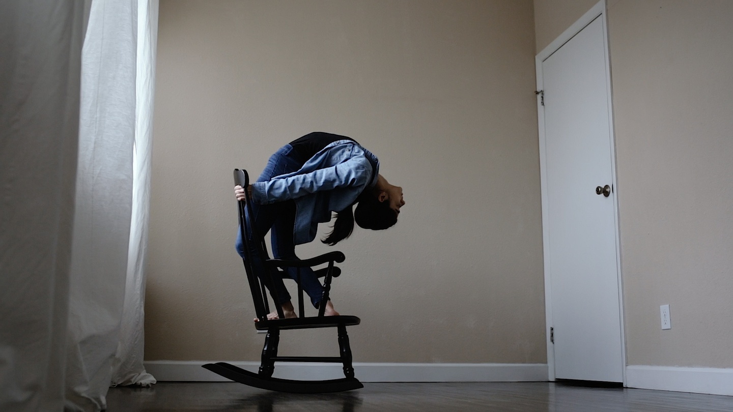 Person in a jeans shirt and leggings stands on a wooden rocking chair in an empty bedroom. They grip the back of the chair and bend all the way backwards.