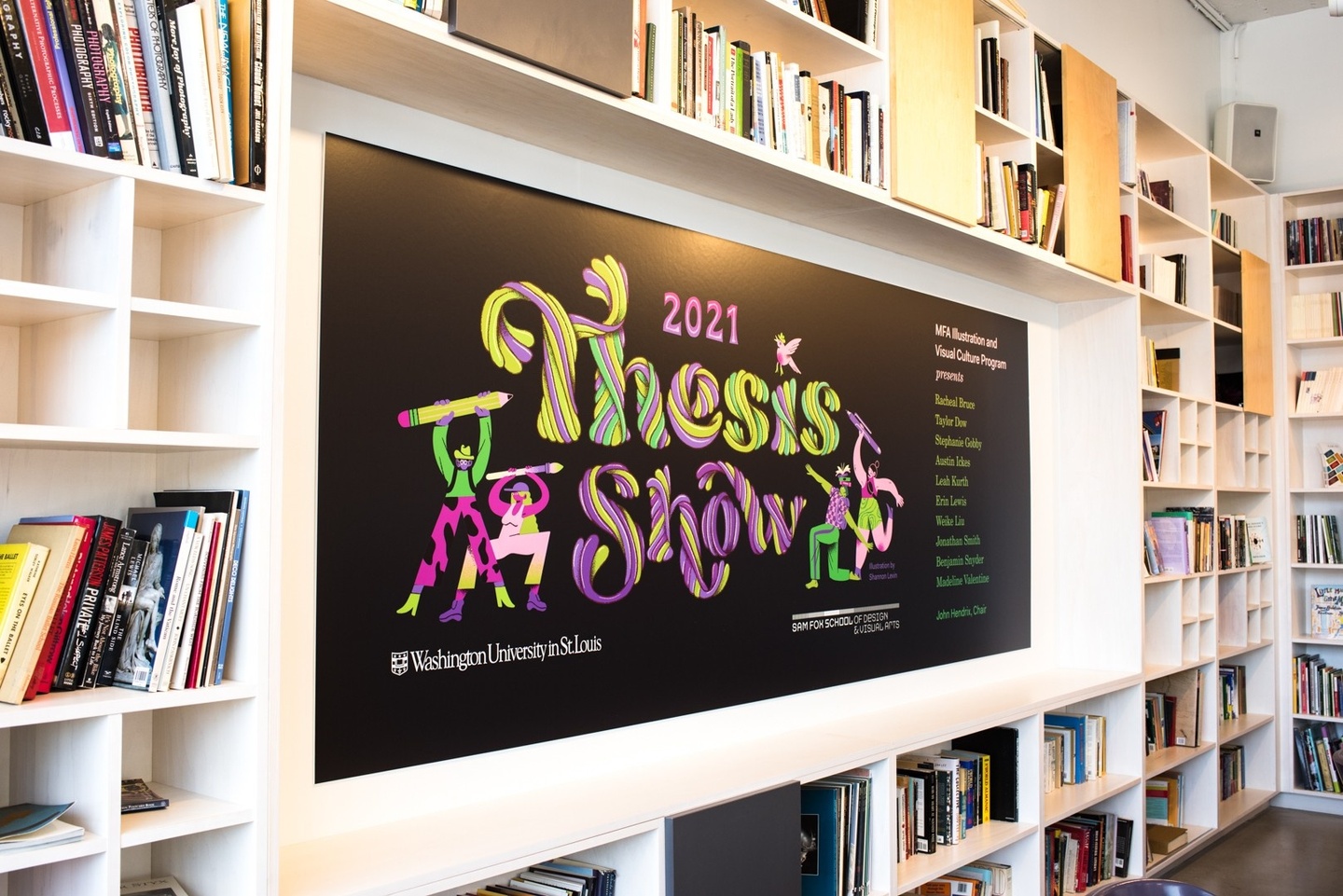 White bookcase wall with an inset frame displaying a black vinyl poster that reads "2021 Thesis Show" in green and purple bubbly letters.