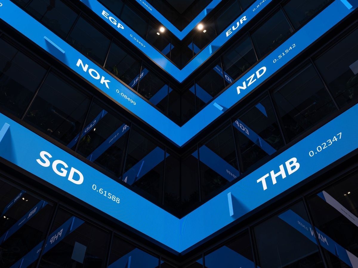 Photograph looking up at four LED screens at the interior intersection of two walls, showing a blue background and information about worldwide currencies