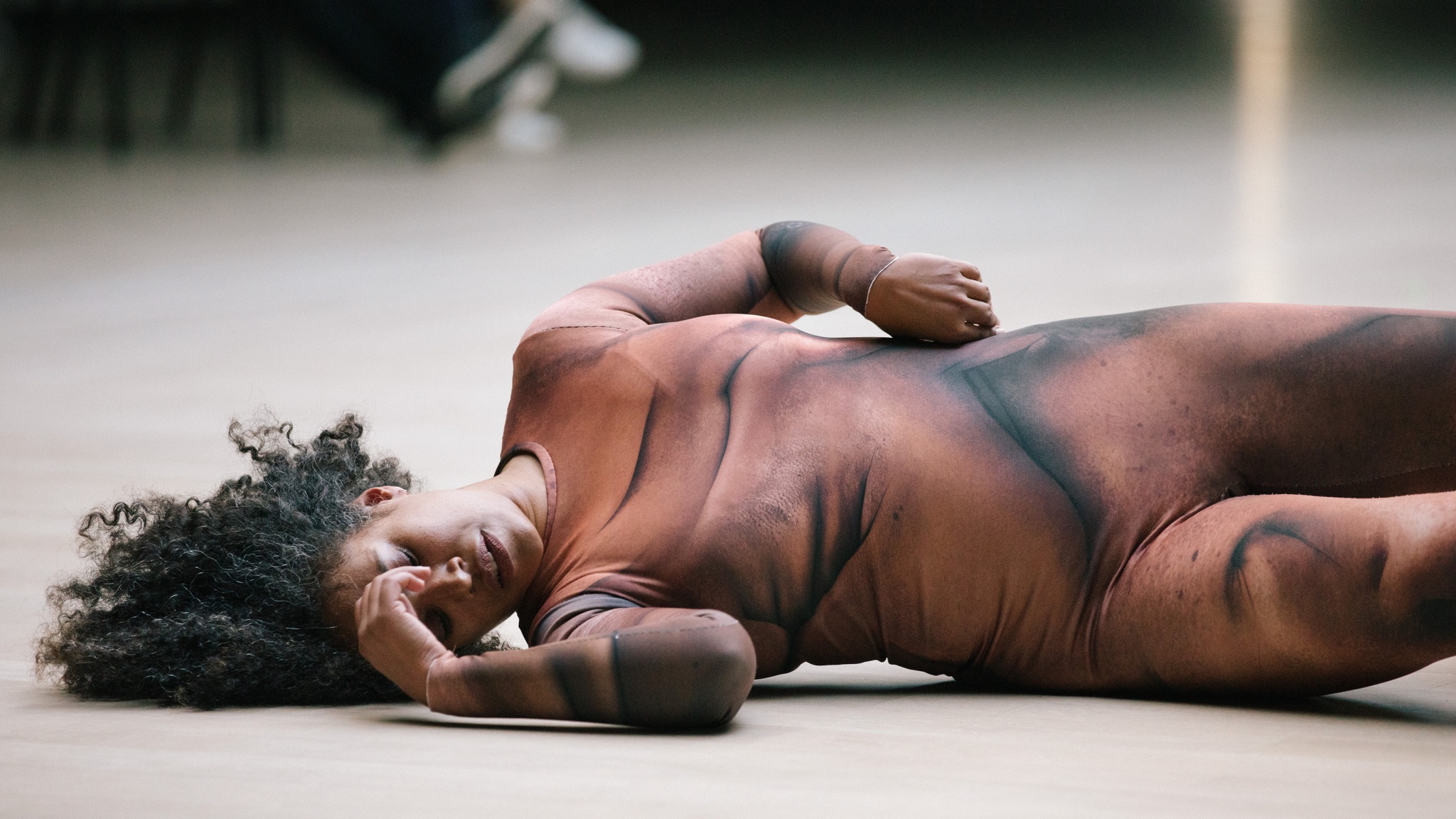 A dancer in a body suited printed with a light and dark brown design lies on the floor with one hand resting on her forehead