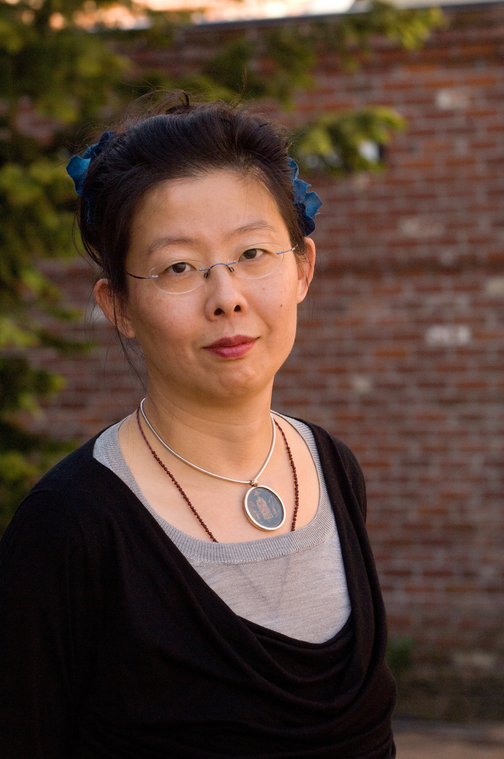 Costume designher Anita Yavich standing in front of a green tree and brick wall. She wears glasses a necklace with a large round medallion and a black deep-v neck shirt over a lighter gray shirt with a higher neck