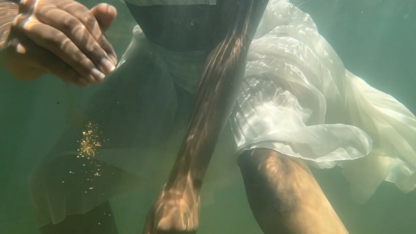 A still from Carolina Caycedo’s video shows a person underwater, wearing a translucent white garment. Their hands, torso, and legs are partially visible through the green-blue water, with strands of long hair drifting across the frame.