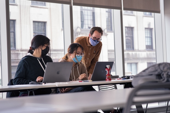 Instructor looks over the shoulders of two students with laptops at a conference table. 