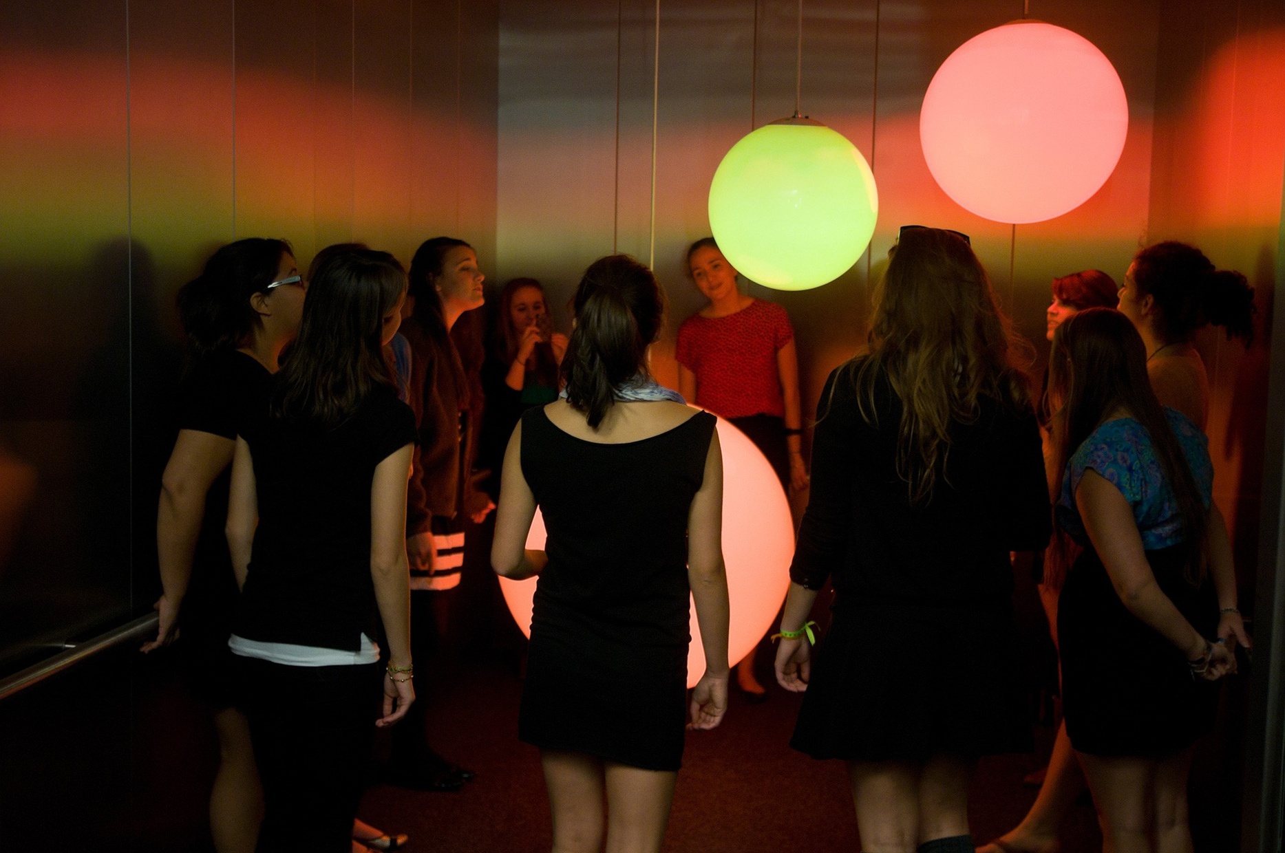 A group of people crowd in an elevator to look at three, large glowing spheres hanging from the ceiling at different heights. Two of the spheres glow red and one glows yellow.